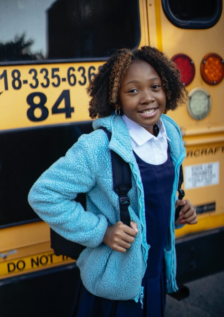 A girl with a backpack smiling in front of a bus on a school trip.